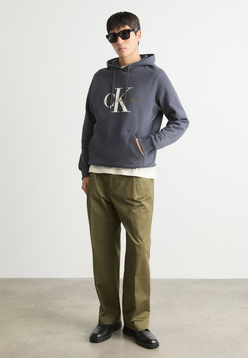 Sudadera con capucha gris con el logo "CK" y el texto "Calvin Klein Jeans", combinada con pantalones verdes oliva y zapatos negros.