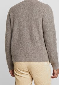 Beige stickad tröja med ribbat mönster, rund halsringning och långa ärmar, tillsammans med krämfärgade kordfläktbyxor.