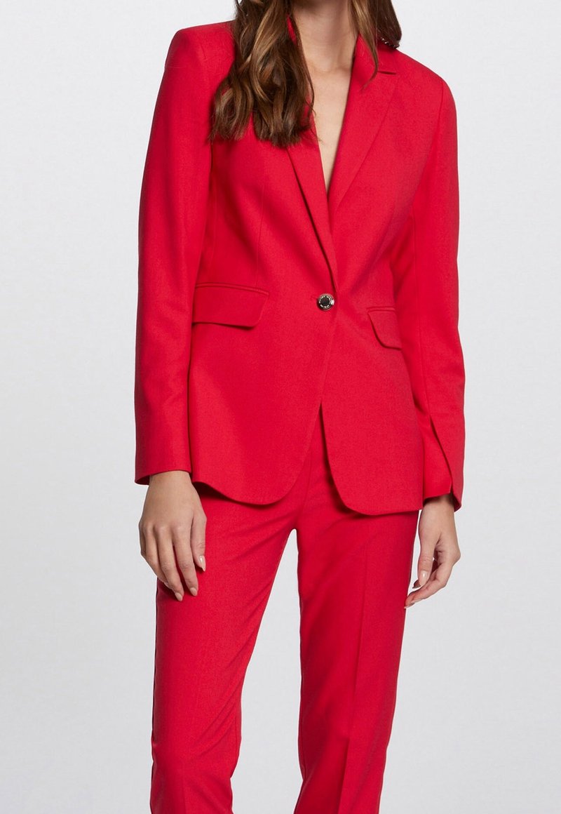 Femme portant un blazer rouge sur mesure et un pantalon rouge assorti, debout devant un fond clair uni.