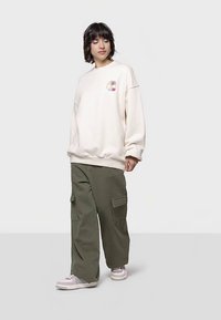 Kremowa, oversize'owa bluza z grafiką na lewej piersi, zestawiona z zielonymi spodniami typu cargo i jasnymi sneakersami.