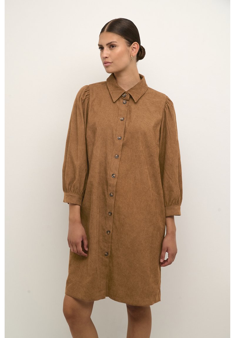 Robe chemise en velours côtelé marron avec un col, de grands boutons sur le devant, des manches bouffantes longues et une coupe décontractée. Texture douce et longueur genoux.