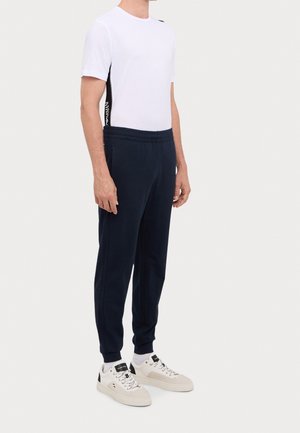 Homme portant un t-shirt blanc à manches courtes avec des bandes latérales noires, un pantalon de jogging bleu marine, des chaussettes blanches et des baskets blanches, debout devant un fond blanc.