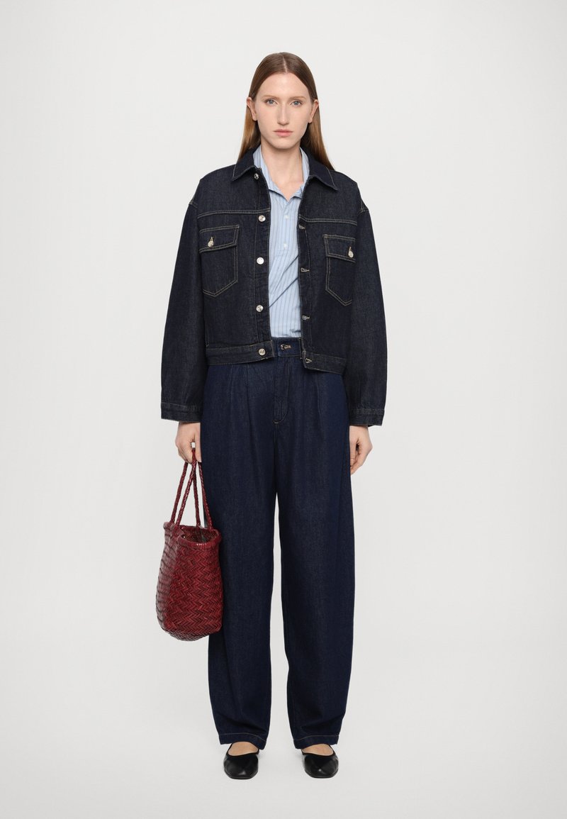 Giacca cropped in denim scuro con due tasche sul petto, abbinata a pantaloni larghi in denim scuro. Tieni una borsa intrecciata rossa e indossi scarpe nere flat. Sotto, una camicia a righe.