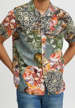 Koszula na krótki rękaw z guzikami, ozdobiona wielokolorowymi geometrycznymi i kwiatowymi wzorami w stylu patchworku, noszona przez osobę z rękami w kieszeniach.