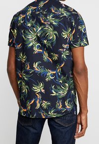 Kortærmet skjorte lavet af navyblåt stof med et livligt tropisk bladmønster i grønne, orange og pink nuancer. Lige kant.