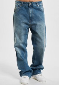 Blå denimjeans med løs passform, som har fem lommer, glidelås og knappelukking, samt diskret falming over hele buksen. Kombinert med hvite sneakers.