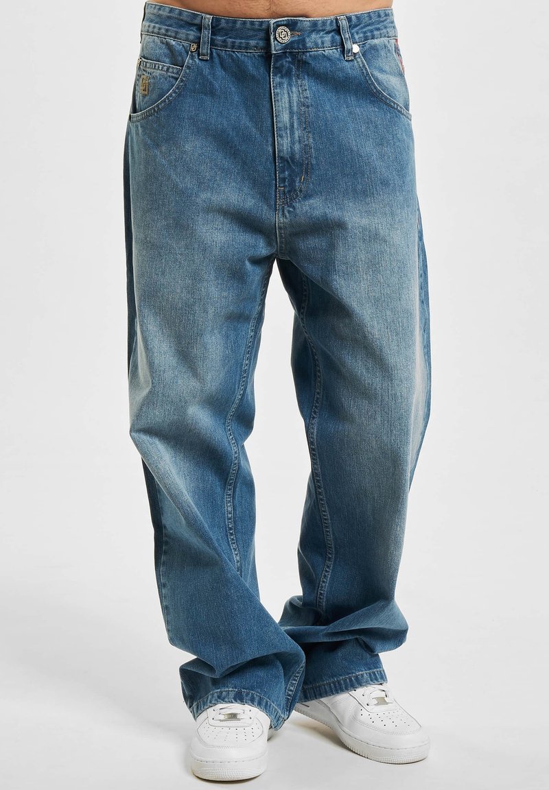 Blå denimjeans med løs passform, som har fem lommer, glidelås og knappelukking, samt diskret falming over hele buksen. Kombinert med hvite sneakers.