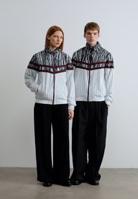 Λευκά μπουφάν με φερμουάρ και υφή με σκούρες οριζόντιες ρίγες, συνδυασμένα με φαρδιά μαύρα παντελόνια. Λείο, σύγχρονο σχέδιο.