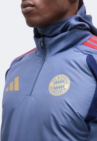 Κοντινό πλάνο ενός ατόμου που φοράει μπλε αθλητική μπλούζα FC Bayern Munich με φερμουάρ στο ένα τέταρτο και κόκκινες ρίγες στους ώμους.