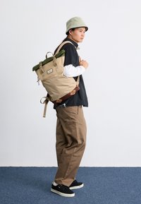 Zaino beige arrotolabile con dettagli verdi, spallacci regolabili e una tasca frontale. Indossato sopra una maglietta nera con pantaloni beige e scarpe nere.