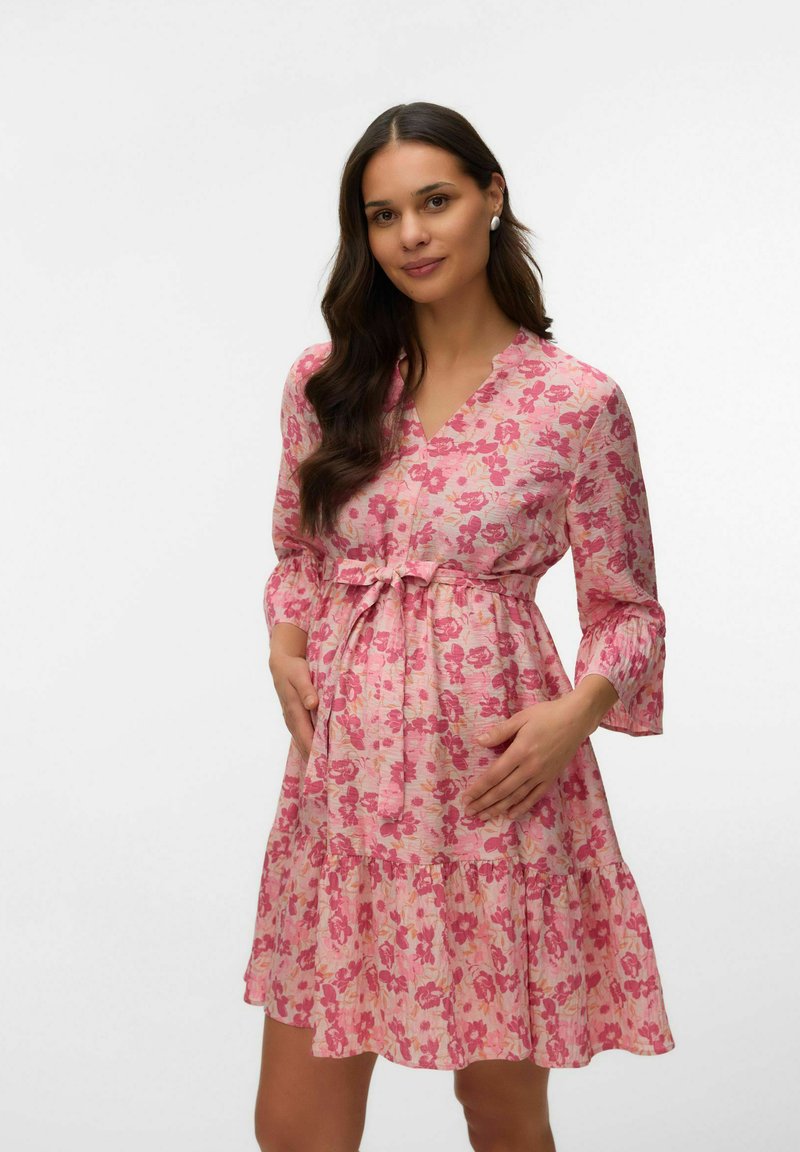 Moteris su ilgais tamsiais plaukais, dėvinti rožinę gėlėtą kelio ilgio suknelę, švelniai laikanti savo nėščio pilvuko sritį, stovinti priešais paprastą baltą foną.