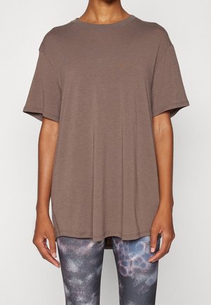 Person wearing an oversized brown short-sleeve shirt paired with black and blue patterned leggings, standing against a plain background.