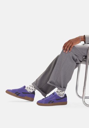 Fioletowe zamszowe trampki z czarnymi akcentami, gumową podeszwą w kolorze naturalnym oraz białymi skarpetkami w wzory, zestawione ze szarymi spodniami, siedzące na krześle.