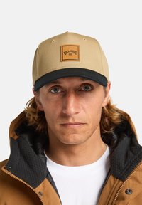 Casquette de baseball beige avec une visière noire et un patch en cuir marron arborant le logo de la marque. Portée avec une veste marron et une chemise blanche.