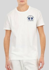 T-shirt blanc à manches courtes avec un logo "La Martina" bleu marine représentant deux joueurs de polo à cheval sur la poitrine gauche.