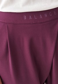 Bordo sportske hlače s glatkom teksturom, elastičnim pojasom i ispisanom riječi "BALANS" duž gornjeg dijela.