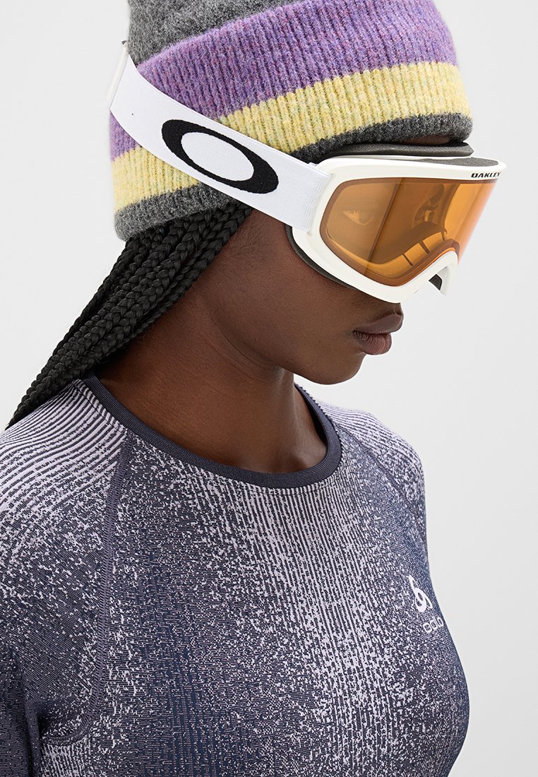 Gafas de esquí con lentes ámbar y marco blanco, llevadas con un gorro de punto gris y púrpura a rayas. Camiseta deportiva azul con estampado.