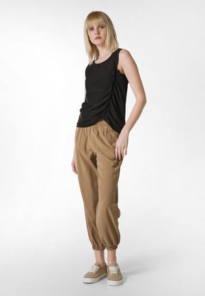 Top senza maniche nera con arricciature laterali, abbinato a pantaloni beige con vita elastica che si restringono alle caviglie. La modella indossa sneakers beige.
