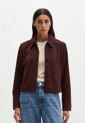 Frau trägt eine dunkelrote, geknöpfte Jacke über einem cremefarbenen Top und blauen, hoch taillierten Jeans und steht vor einem einfarbigen hellen Hintergrund.