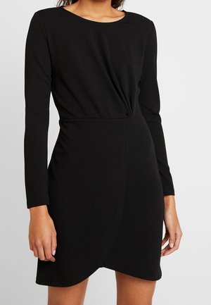 Kobieta w dopasowanej czarnej sukience z długim rękawem, z ozdobnym skrętem w talii i tulipanowym dołem, na jednolitym tle.