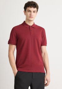 Bordo polo marškiniai su apykakle ir dviem mygtukais, pagaminti iš glotnios medžiagos. Itin subtilus prekės ženklo ženklas ant krūtinės.