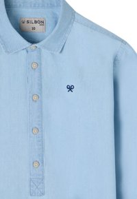 Camisa de color azul claro con cuello y una tapeta de botones, con botones en color crema y un pequeño logo de tijeras en navy en el lado izquierdo del pecho.