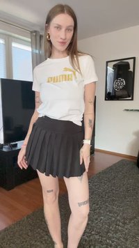 Camiseta blanca con el logo dorado de "PUMA", combinada con una falda negra plisada. Configuración interior con suelo de madera y una alfombra oscura.
