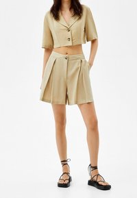 Beige kort kavaj med korta ärmar, knappstängning och framfållade shorts. Kombinerad med svarta remsandaler. Slät tygkänsla.