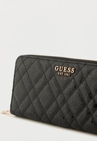 Czarny pikowany portfel wykonany z teksturowanego materiału, z złotym logo "GUESS EST 1981" oraz zamkiem błyskawicznym. Wykończony gładką lamówką.