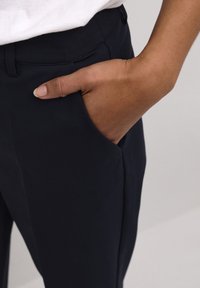 Pantalon extensible bleu marine avec une texture lisse, doté d'une poche latérale et d'une ceinture. Main partiellement visible dans la poche.