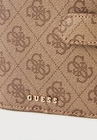 Hnědá texturovaná peněženka s jemným vzorem loga, na přední straně se zlatou destičkou značky "GUESS". Kompaktní obdélníkový tvar.
