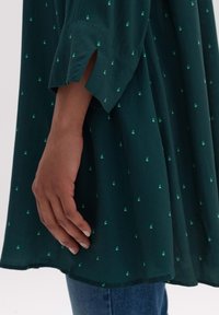 Tunika verde scuro con sottili disegni di foglie verdi, in tessuto morbido, con polsini a spacco e vestibilità rilassata, indossata sopra un paio di jeans azzurri.