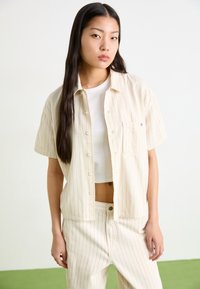 Camicia a maniche corte, colore crema con sottili righe rosse e tasca sul petto, abbinata a pantaloni a righe coordinati. Tessuto liscio, vestibilità casual.