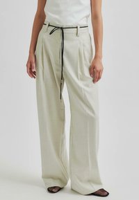 Pantaloni larghi beige chiaro con vita alta, pieghe frontali e un laccio nero. Il tessuto appare liscio con una leggera lucentezza.