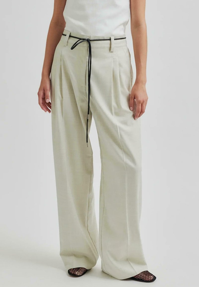 Pantaloni larghi beige chiaro con vita alta, pieghe frontali e un laccio nero. Il tessuto appare liscio con una leggera lucentezza.