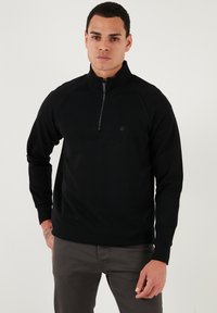 Melna sporta krekla ar ceturtdaļas rāvējslēdzēju, raglana piedurknēm un mazu logotipu kreisajā pusē. Mīksts, strukturēts materiāls un pieguloša piegriezuma dizains.