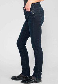 Jeans de mezclilla de un azul oscuro con un corte ajustado, cinco bolsillos y un detalle de costura en contraste. Usados con mocasines negros brillantes.