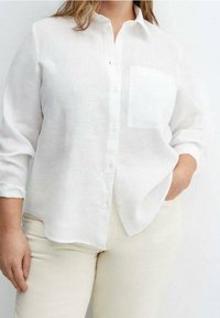 Mujer vestida con una camisa blanca de botones de manga larga con un bolsillo en el pecho y pantalones beige claros, una mano en el bolsillo, frente a un fondo sencillo.