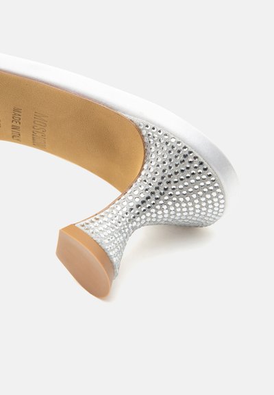 Chaussure à talons hauts argentée avec des ornements en strass sur le talon et une semelle beige, photographiée sur fond blanc.