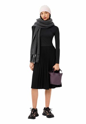 Femme portant une robe noire à manches longues, une écharpe grise, un bonnet rose, des baskets noires, tenant un petit sac à main rayé violet.