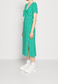 Zielona kwiatowa sukienka midi z dekoltem w serek i wiązaniem, z krótkimi bufkami i przednim rozcięciem, zestawiona z białymi masywnymi trampkami.