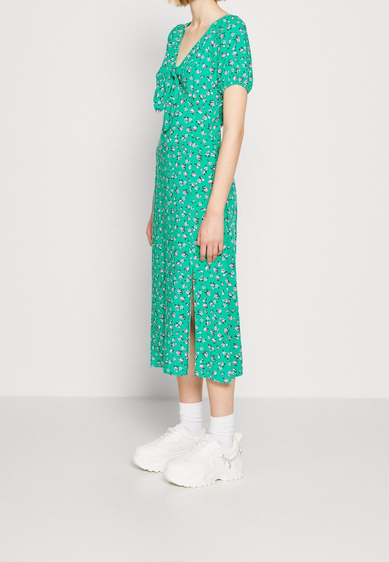 Zielona kwiatowa sukienka midi z dekoltem w serek i wiązaniem, z krótkimi bufkami i przednim rozcięciem, zestawiona z białymi masywnymi trampkami.