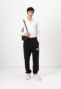 Zwarte trainingsbroek met een elastische tailleband, met logo detail. Witte langemouwen shirt, grijze schoudertas en witte sneakers.