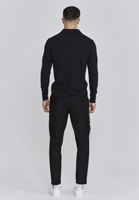 Camisa polo negra de manga larga para hombre con cuello, combinada con pantalones cargo negros que cuentan con bolsillos laterales y zapatillas blancas, todo sobre un fondo neutro.