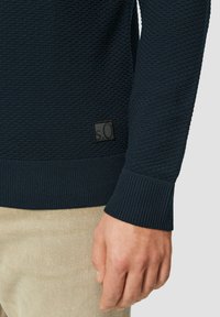 Marineblauwe gebreide sweater met ribbels aan de mouwen en onderrand, met een vierkante stoffen logo patch aan de onderzijde. Beige broek zichtbaar daaronder.