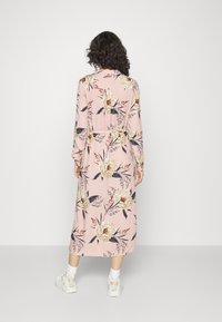 Abito lungo a maniche lunghe in rosa pallido, con grandi fiori crema e blu navy, vita cinta e design fino alla caviglia. Indossato con scarpe bianche.