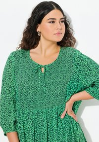 Abito verde con macchie leopardate nere, colletto arrotondato, dettaglio con fiocco e maniche larghe. Tessuto leggero con un corpo testurizzato e una gonna fluida.
