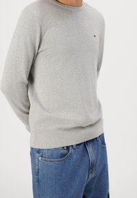 Homme portant un pull à manches longues gris clair et un jean en denim bleu, debout les mains derrière le dos devant un fond uni.