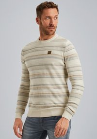 Cream striped sweater with various horizontal lines in beige, navy, and light brown. Featuring a round neck and a small branded patch on the chest.