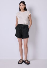 Camicia beige a maniche corte, taglio rilassato; pantaloni corti neri in lino con vita elastica e tasche laterali; sandali neri con lacci e dettagli argentati.